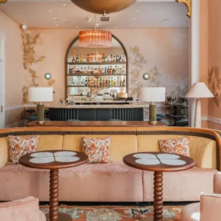 Pastel pink lounge with a curved peach sofa, floral cushions, and two round tables in front of a backlit bar.