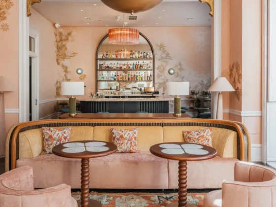 Pastel pink lounge with a curved peach sofa, floral cushions, and two round tables in front of a backlit bar.
