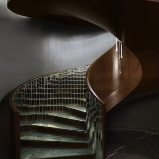 Curved wooden spiral staircase with glass railing, descending in a dimly lit interior.