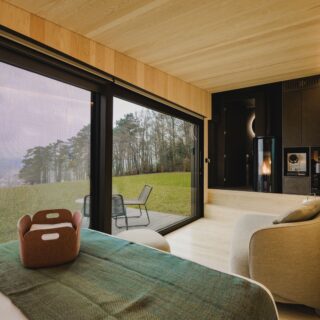 Modern bedroom with floor-to-ceiling glass doors opening to a grassy outdoor deck and chairs; warm wood paneling and a cozy seating area next to a fireplace.