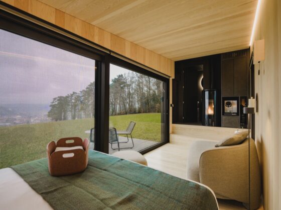 Modern bedroom with floor-to-ceiling glass doors opening to a grassy outdoor deck and chairs; warm wood paneling and a cozy seating area next to a fireplace.