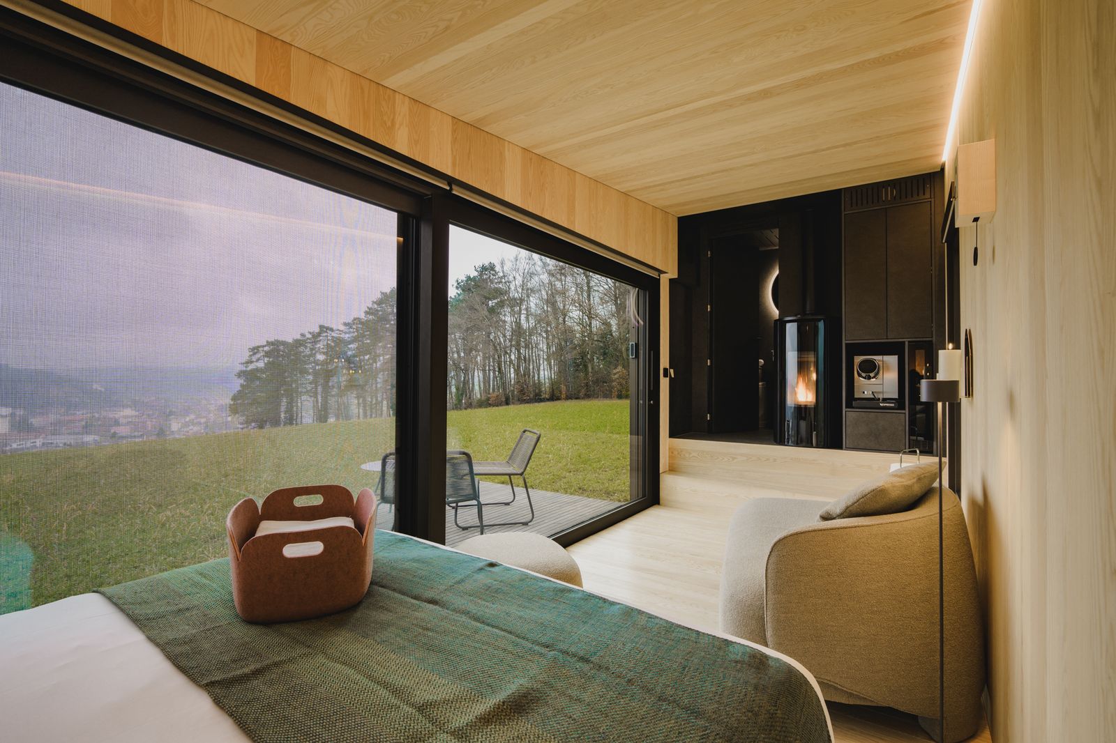 Modern bedroom with floor-to-ceiling glass doors opening to a grassy outdoor deck and chairs; warm wood paneling and a cozy seating area next to a fireplace.
