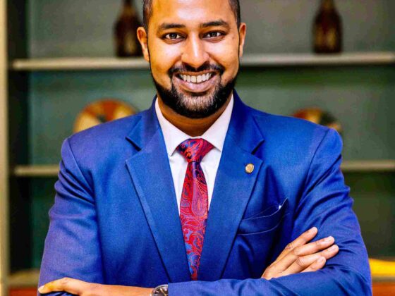 Smiling man in a blue suit with a red patterned tie, arms crossed, in an office setting.