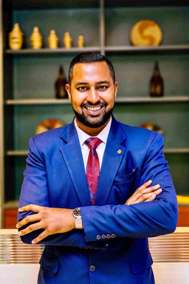 Smiling man in a blue suit with a red patterned tie, arms crossed, in an office setting.