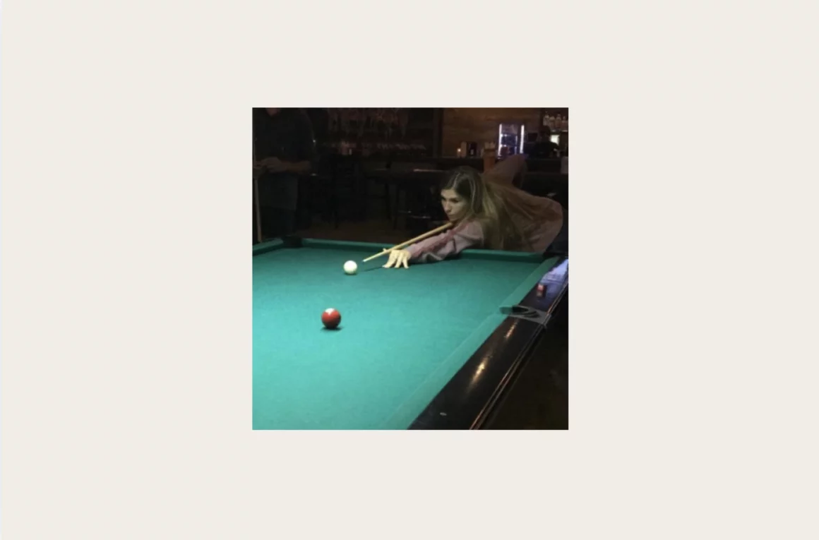 Woman leaning over a green pool table, aiming a cue at the white cue ball in a dim bar setting.