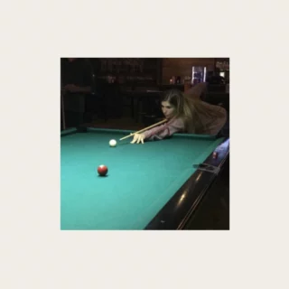 Woman leaning over a green pool table, aiming a cue at the white cue ball in a dim bar setting.
