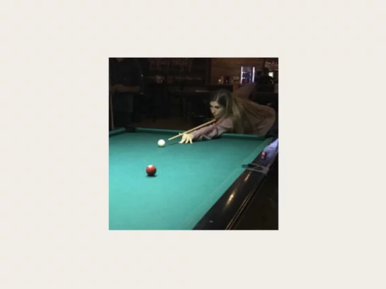 Woman leaning over a green pool table, aiming a cue at the white cue ball in a dim bar setting.