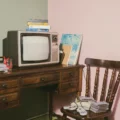 Retro living-room scene with a vintage CRT TV on a wooden desk, stacks of VHS tapes, and old game controllers on a chair nearby, against a pink wall.