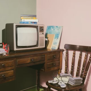 Retro living-room scene with a vintage CRT TV on a wooden desk, stacks of VHS tapes, and old game controllers on a chair nearby, against a pink wall.