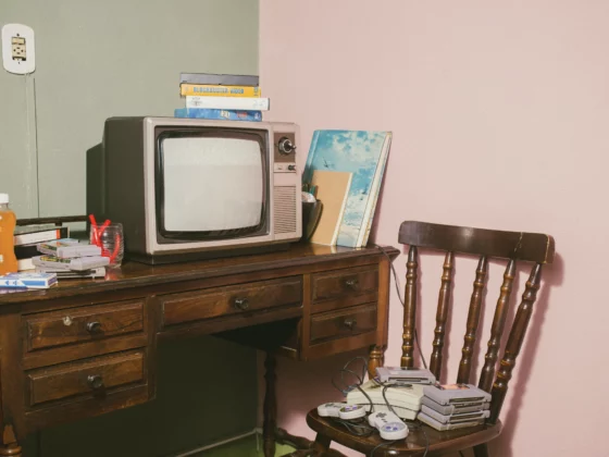 Retro living-room scene with a vintage CRT TV on a wooden desk, stacks of VHS tapes, and old game controllers on a chair nearby, against a pink wall.