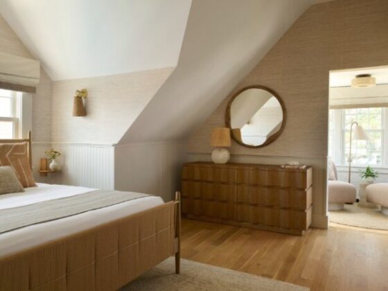 Cozy neutral bedroom with a sloped ceiling, bed, and wooden dresser with a round mirror against beige walls.