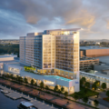 A modern waterfront hotel complex with a tall glass facade, outdoor pool, and marina along a promenade at dusk/light.