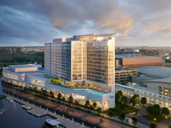 A modern waterfront hotel complex with a tall glass facade, outdoor pool, and marina along a promenade at dusk/light.