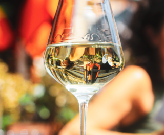 Close-up of a wine glass on an outdoor dining table, with a blurred person reading a menu in the background.