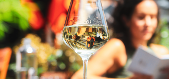 Close-up of a wine glass on an outdoor dining table, with a blurred person reading a menu in the background.