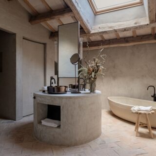 Rustic bathroom with a curved concrete island, tall mirror, and a dried floral arrangement; skylight above and a freestanding tub on the right.