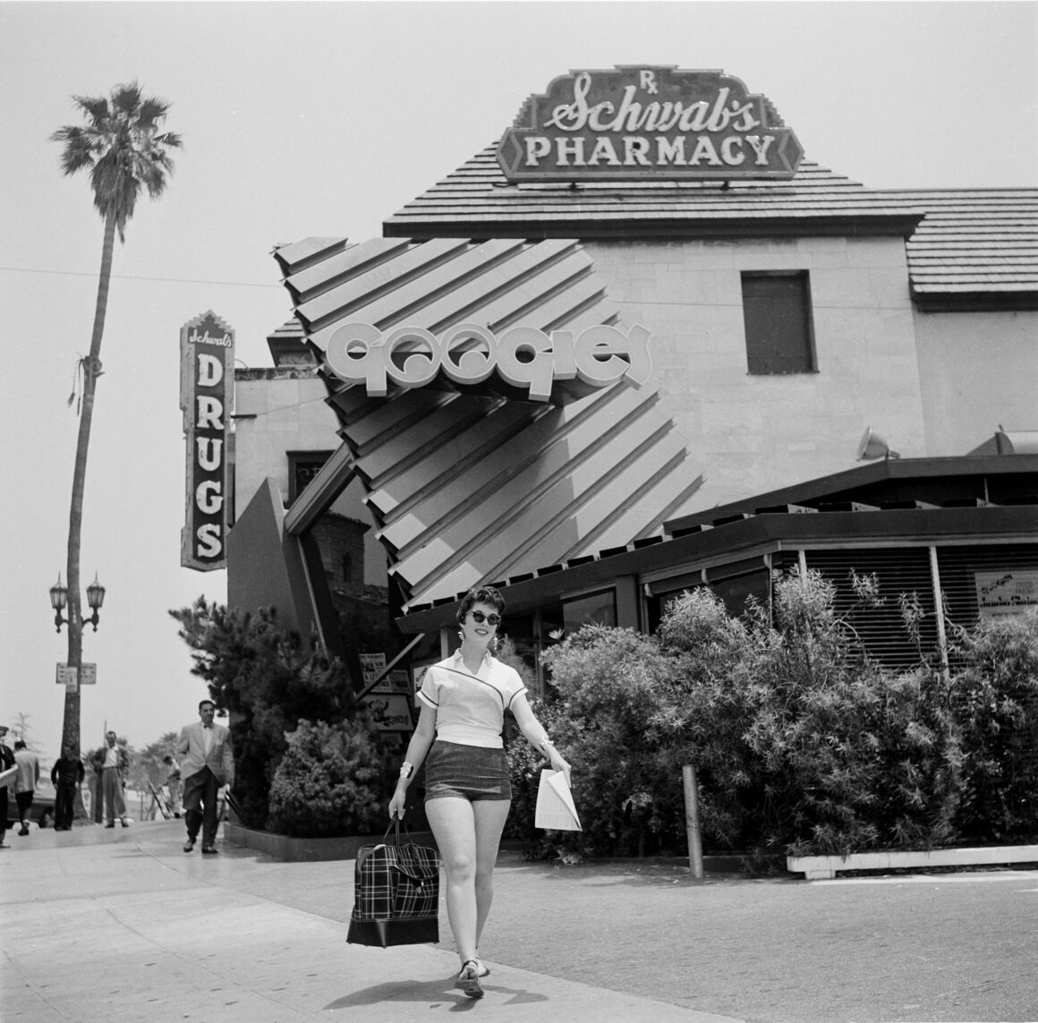 Woman walking on a sunny street pulling a large plaid shopping bag; retro storefronts and a Schwab's Pharmacy sign in the background.