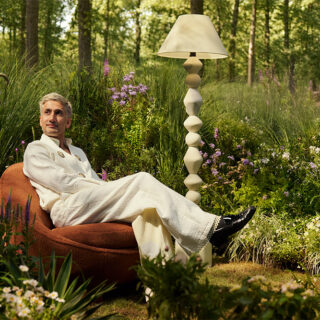 Man in a white suit lounging in a cushioned brown chair in a lush garden, with a tall decorative lamp nearby.