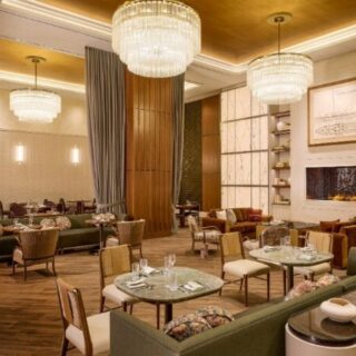 Luxurious hotel lounge with green sofas, round tables, and crystal chandeliers near a fireplace.