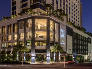 Modern multi-story shopping complex at dusk with illuminated storefronts and palm trees along the street.