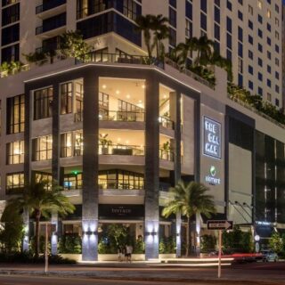 Modern multi-story shopping complex at dusk with illuminated storefronts and palm trees along the street.