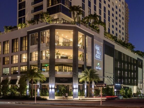 Modern multi-story shopping complex at dusk with illuminated storefronts and palm trees along the street.