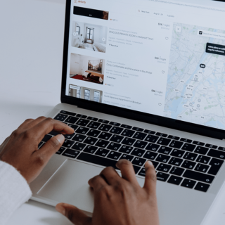 Person typing on a laptop showing Airbnb search results with listing thumbnails and a map on the screen, browsing accommodations in New York City area.