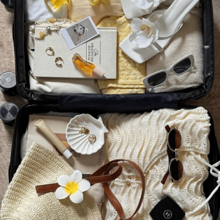 Open suitcase filled with summer travel essentials: white floral sandals, sunglasses, jewelry, perfume, and a straw bag.
