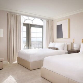 Hotel room with two white beds, beige headboards, and balcony doors framed by light curtains.