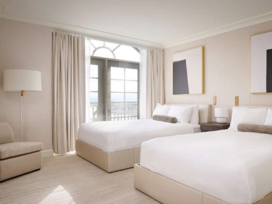 Hotel room with two white beds, beige headboards, and balcony doors framed by light curtains.