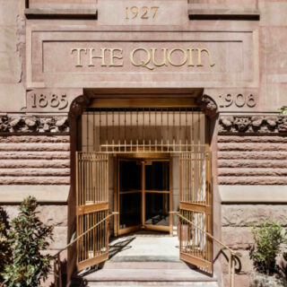 Stone building entrance labeled THE QUOIN with 1927 inscription; ornate doorway opens to gold gates and a recessed lobby.