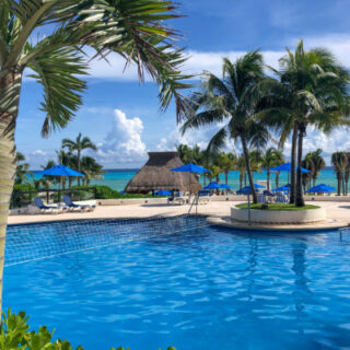 Poolside at a tropical resort with palm trees, lounge chairs under blue umbrellas, and the ocean in the background.