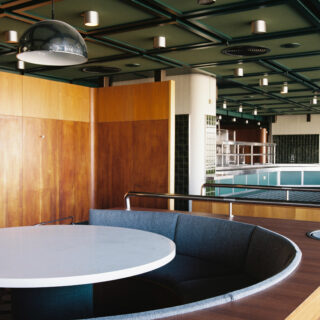 Interior view of a modern lounge with a large white round table and curved blue booth seating against wooden panels. The ceiling has green beams and pendant lights.