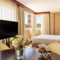 Hotel suite with a round table in the foreground, a vase of white flowers, a flat-screen TV, and a bed by large windows with a city view outside.