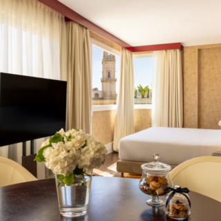 Hotel suite with a round table in the foreground, a vase of white flowers, a flat-screen TV, and a bed by large windows with a city view outside.