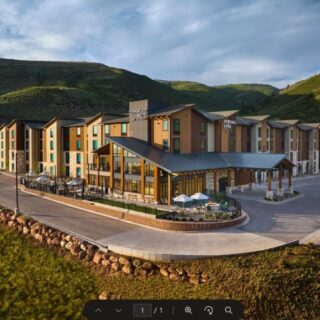 A modern lodge-style hotel with outdoor dining area and umbrellas, set against green hillside and blue sky.