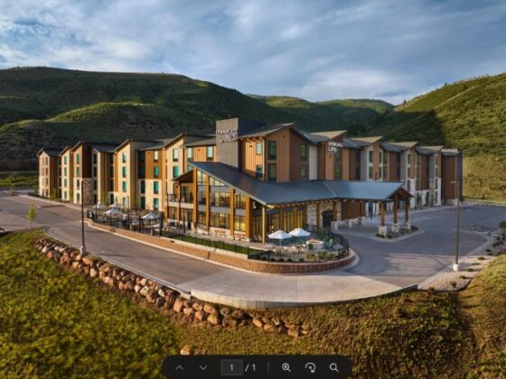 A modern lodge-style hotel with outdoor dining area and umbrellas, set against green hillside and blue sky.