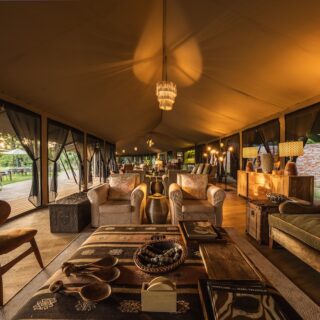 Luxurious tent lounge with beige sofas, plush chairs, wooden coffee tables, and warm amber lighting; visible patio outside the canopy.