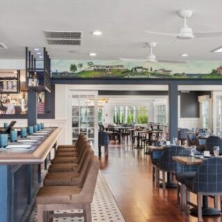 Open-plan restaurant interior with a blue bar on the left, brown bar stools, and blue upholstered dining chairs around wooden tables.”],