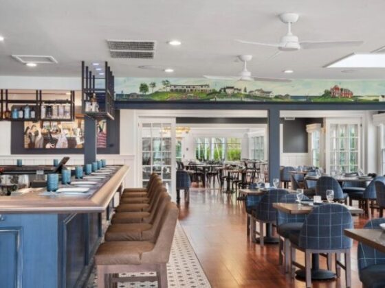 Open-plan restaurant interior with a blue bar on the left, brown bar stools, and blue upholstered dining chairs around wooden tables.”],