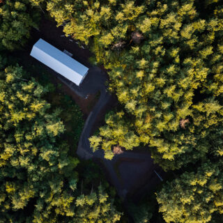 Aerial view of a long rectangular white building in a dense evergreen forest, with a curved dirt road around it.