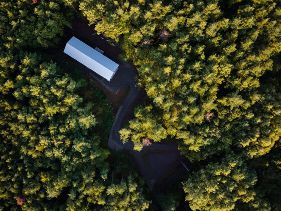 Aerial view of a long rectangular white building in a dense evergreen forest, with a curved dirt road around it.