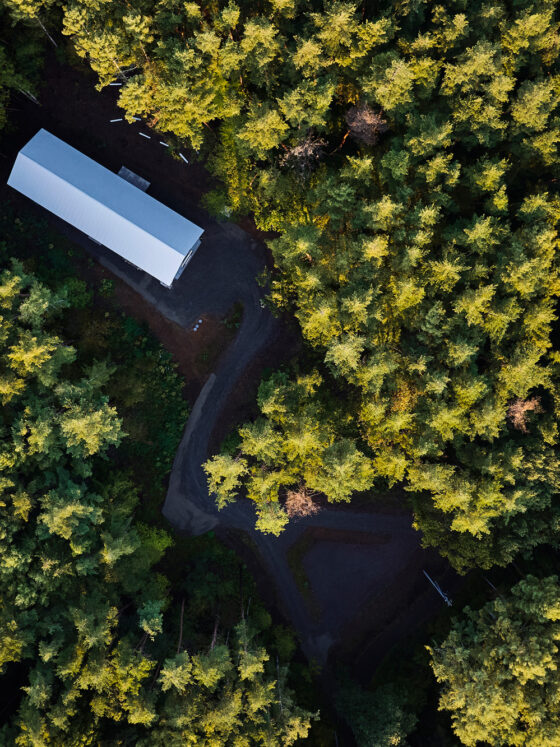 Aerial view of a long rectangular white building in a dense evergreen forest, with a curved dirt road around it.