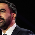 Close-up of a bearded man in a dark suit speaking on stage, looking slightly to the side.