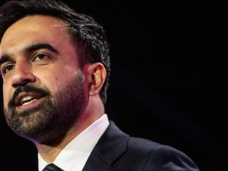 Close-up of a bearded man in a dark suit speaking on stage, looking slightly to the side.