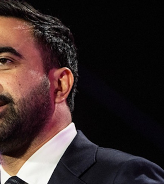 Close-up of a bearded man in a dark suit speaking on stage, looking slightly to the side.