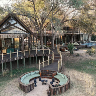 Aerial view of a woodland resort with a circular wooden lounge and teal-cushioned seating around a fire pit under trees on a sandy ground.