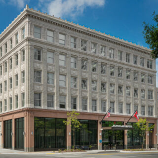 Four-story classic brick-and-stone building with glass storefronts on the ground level and flags at the entrance on a city street.