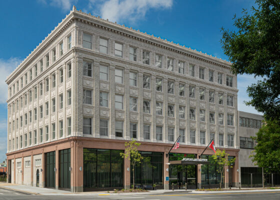 Four-story classic brick-and-stone building with glass storefronts on the ground level and flags at the entrance on a city street.