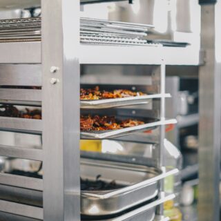 Stainless steel rack with multiple baking trays in a commercial kitchen, trays filled with roasted foods